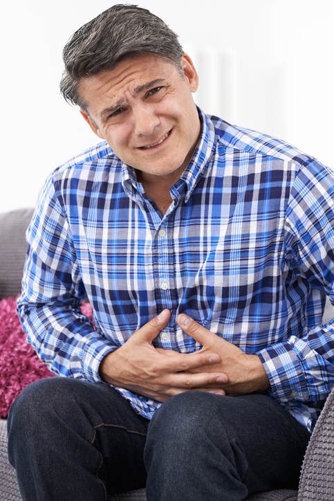 IBS - muž má bolesti břicha, křeče, drží se za břicho, modrá kostkovaná košile, sedí na pohovce