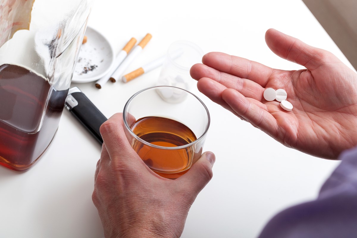Alkohol, kouření a léky. Člověk drží sklenici hnědého alkoholu a léky v pravé ruce. Jako nejčastější příčiny chronické pankreatitidy