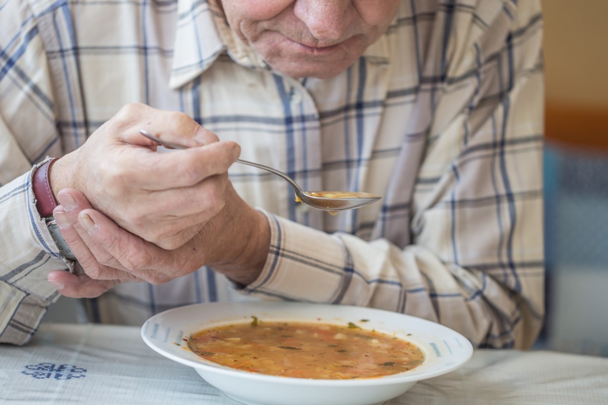 Muž jí polévku a drží si ruku proti třesu při Parkinsonově chorobě