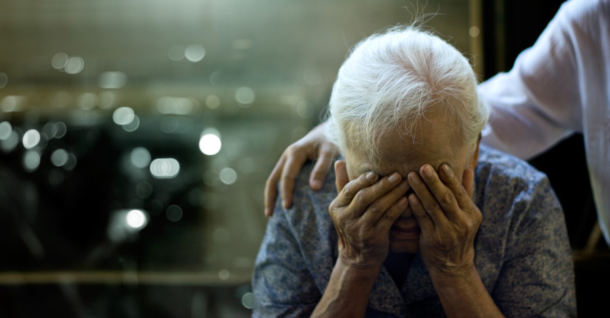 Starší žena má zdravotní problém, hlavu má podepřenou rukama a za rameni ji drží osoba