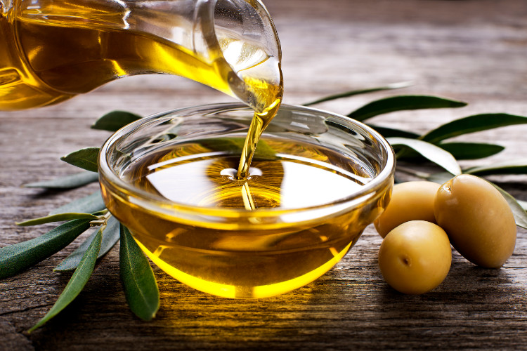 olivový olej někdo lije z číše do misky, vedle je jsou olivy a listy z olivovníku