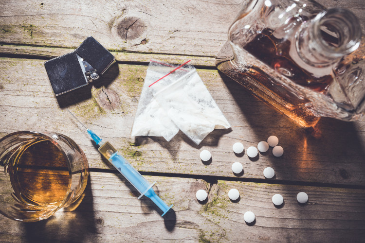 na dřevěném stole je skleněná sklenice a láhev s alkoholem, rozsypané léky, injekční stříkačka, sáčky s drogami a zapalovač