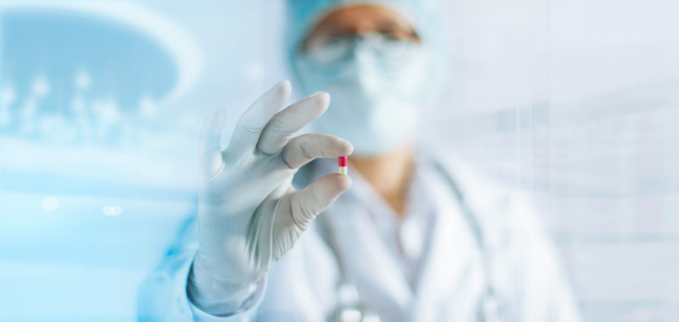 doktorka v bílé uniformě s rouškou na obličeji drží v ruce pilulku