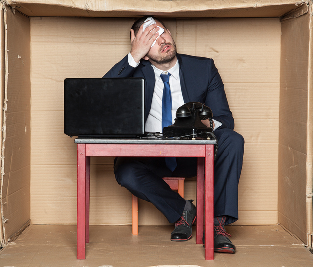 Byznysmen sedí za stolem v krabici, stísněné prostory, pracovní stres a zvýšená zátěž, počítač a telefon, muž si utírá čelo
