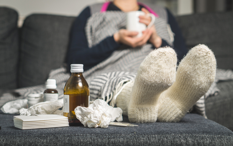 nemocná žena leží v posteli, má teplé ponožky a vedle sebe pilulky a sirupy