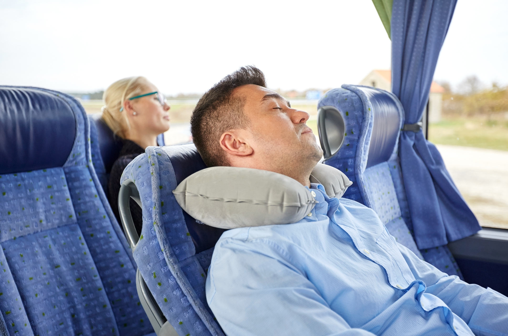 Dva cestující, spící v autobuse, muž má polštář. Sedadla, venku přes okna vidět stromy