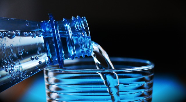 Voda se nalévá s láhve do sklenice. Je vidět pouze hrdlo láhve a vršek sklenice. Pozadí je tmavé a modré.