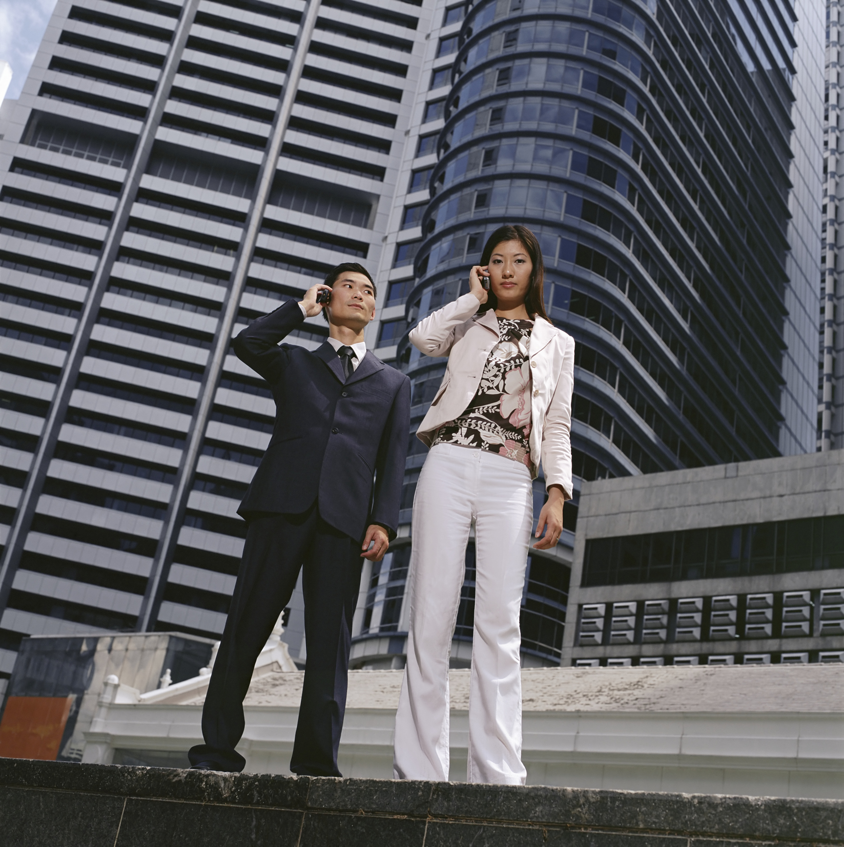 Žena a muž telefonují z mobilů, v pozadí výškové budovy.