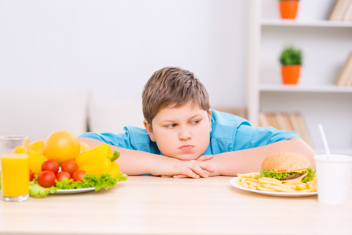 Tlustý kluk mezi dvěma talíři se zdravým a nezdravým jídlem.