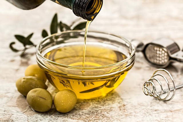 Do skleněné misky se nalévá olivový olej ze skleněné láhve. Kolem misky jsou uloženy zelené olivy.