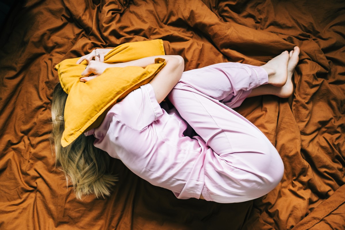 Unavená žena s bolestí leží v posteli a přikrývá si hlavu polštářem.