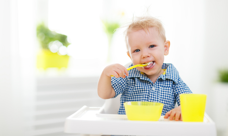 Malé dítě sedí v dětské sedačce, krmí se. Žlutá lžička, pohár a miska