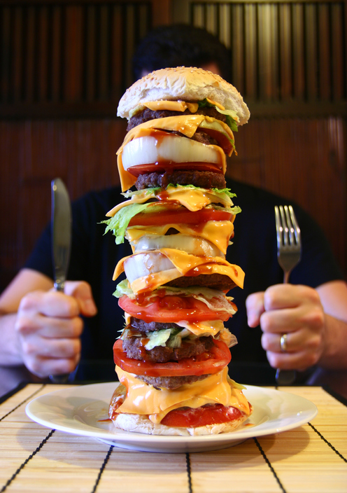 Muž sedí za stolem, na talíři má vysoký hamburger, megaporce